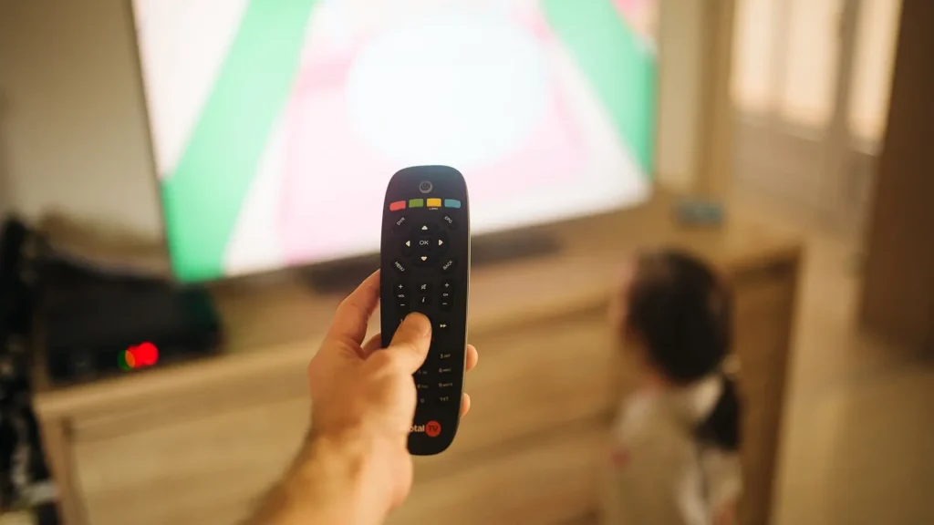 Hand pointing a remote control at a living-room TV while a child watches, the moment IPTV buffering usually gets noticed