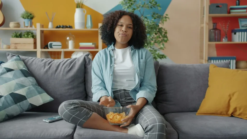 Young woman relaxing on a sofa with a bowl of crisps and a TV remote beside her, ready to watch Kodi on Firestick