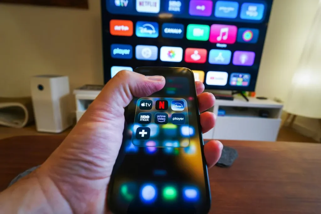 Hand holding a smartphone showing streaming apps in front of a smart TV home screen
