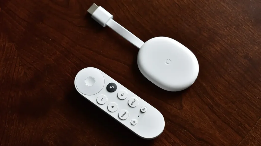 White Chromecast with Google TV streaming device and its remote on a dark wooden table, a typical smart TV box